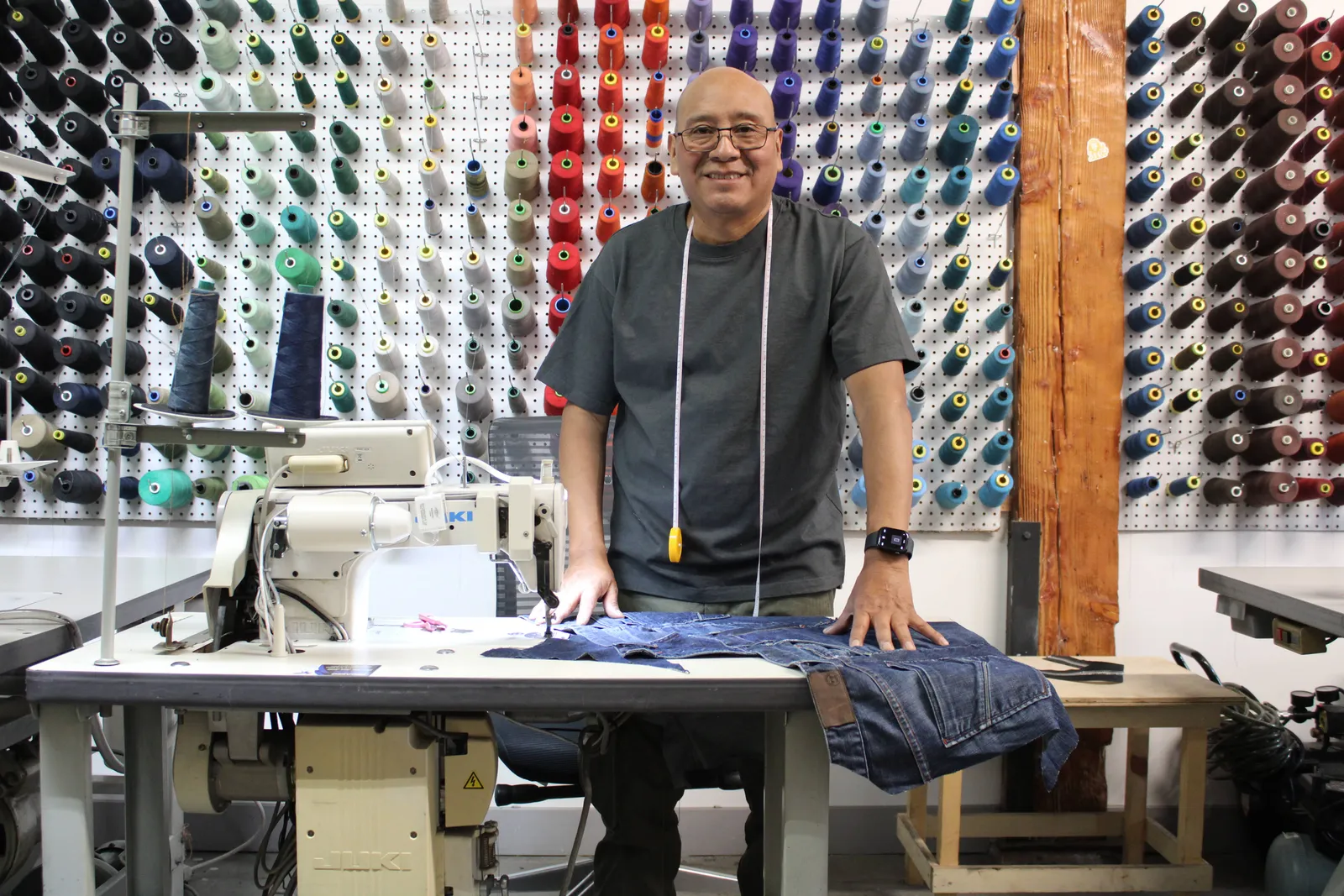 El sastre de JC Atelier ajustando un pantalón de mezclilla en su máquina, frente al muro de conos de hilo del taller